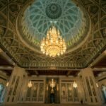 Ornate interior of the Sultan Qaboos Grand Mosque in Muscat, Oman, featuring a large, intricate chandelier hanging from a beautifully patterned dome ceiling. The mosque’s architecture showcases detailed Islamic art and craftsmanship.