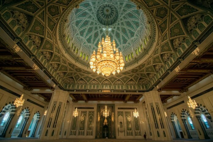 Ornate interior of the Sultan Qaboos Grand Mosque in Muscat, Oman, featuring a large, intricate chandelier hanging from a beautifully patterned dome ceiling. The mosque’s architecture showcases detailed Islamic art and craftsmanship.